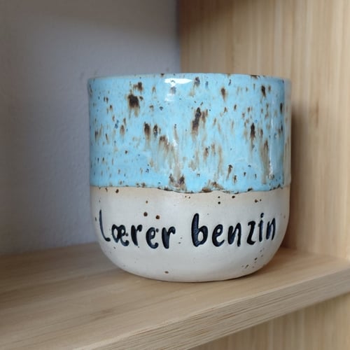 Keramikkop med blå og beige glasur, hvorpå der står "Lærer benzin" i sort tekst, placeret på en lys træhylde. (1)