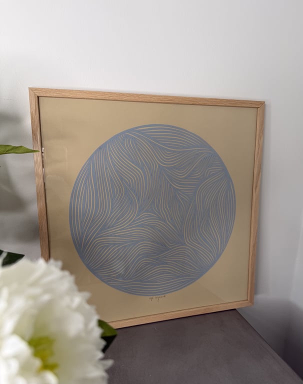 Abstrakt billedkunst med blå cirkel og bølgende linjer i træramme, placeret på gråt bord med hvid blomst i forgrunden.