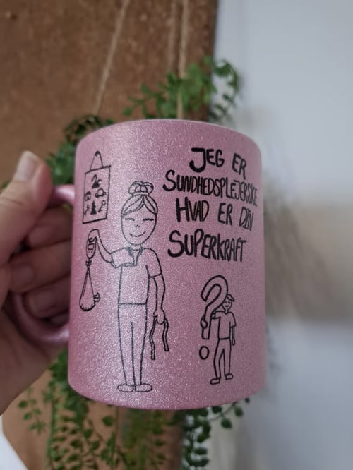 En lys lilla kop med glitrende overflade, der viser en tegning af en kvinde i sundhedsplejerskeuniform og teksten "Jeg er sundhedsplejerske, hvad er din superkraft". En hånd holder koppen, og der er grønne planter i baggrunden. (1)