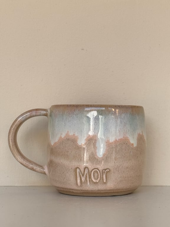 Beige keramikkop med blå og hvide glazurdetaljer og ordet "Mor" præget på siden.