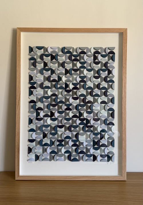 Billede af en indrammet abstrakt kunstplakat med mønster bestående af gentagne buede, halvcirkelformede elementer i blå, grå og sorte nuancer, placeret på en hylde mod en lys væg. (2)