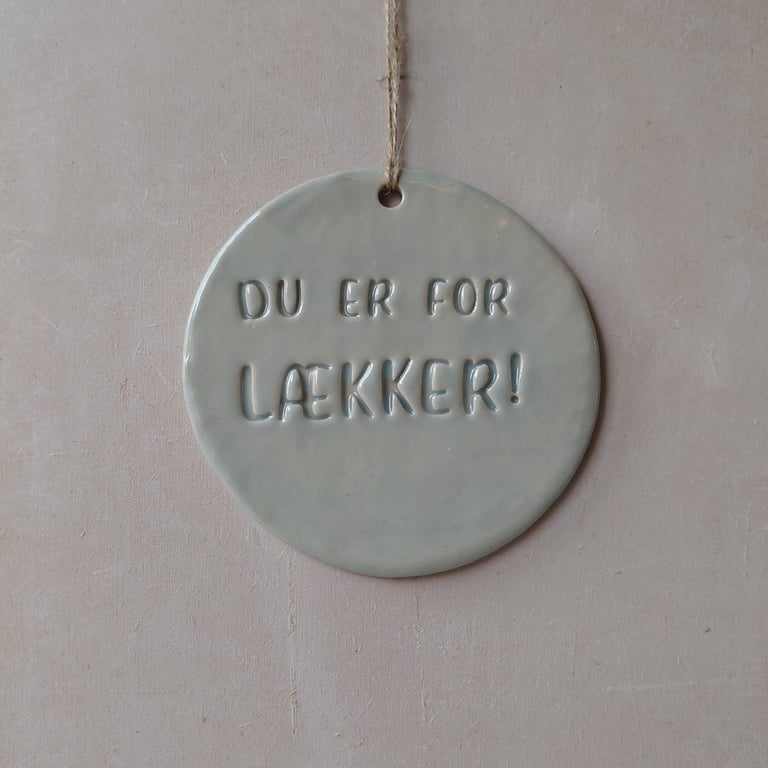 Rundt, gråt keramikskilt med teksten "DU ER FOR LÆKKER!" hængende i en snor mod en beige væg.