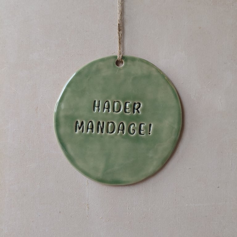 Grøn keramikplade med teksten "HADER MANDAGE!" hængende på en snor mod en beige væg.