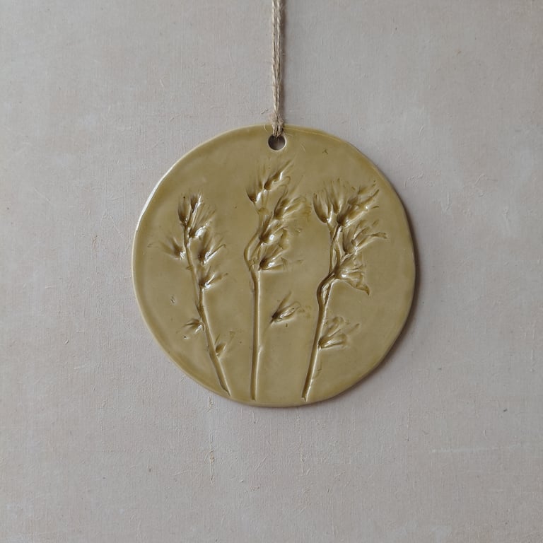 Rund keramisk vægdekoration med aftryk af tre stiliserede planter, hængende i en snor mod en lys væg.