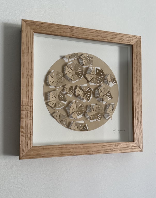 Indrammet papirkunst med tredimensionelle, udskårne blomsterformer i beige og hvide nuancer arrangeret i en cirkel på hvid baggrund, omrammet af en lys træramme.