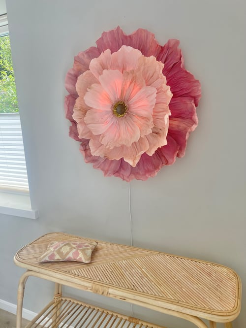 Stor dekorativ blomst i nuancer af lyserød og pink hænger på hvid væg over et lyst bambusbord med en lille pude. (1)