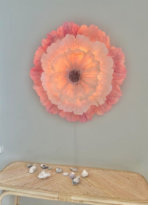 Væglampe formet som en stor, oplyst blomst med rosa og lyserøde kronblade, monteret på en grå væg over et lyst flettet bord med dekorationer. (1)