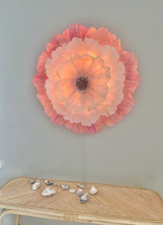 Væglampe formet som en stor, oplyst blomst med rosa og lyserøde kronblade, monteret på en grå væg over et lyst flettet bord med dekorationer.