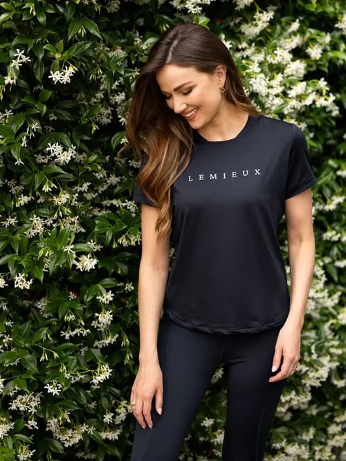 Kvinde iført sort t-shirt med teksten "LEMIEUX" og sorte bukser, stående foran en grøn hæk med hvide blomster, smilende og kigger nedad. (1)