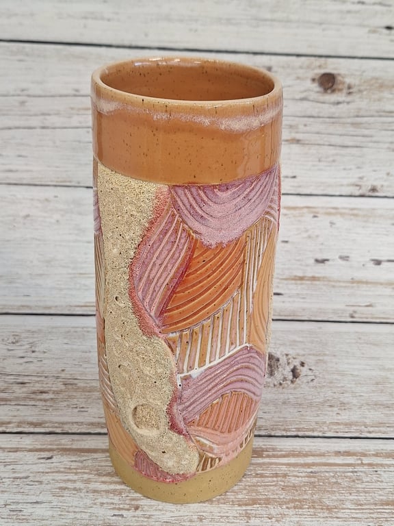 Keramisk cylinderformet vase med abstrakt reliefmønster i rosa, orange og beige toner på en hvid træbaggrund.