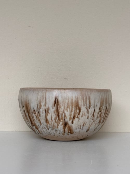 Keramisk skål med brun og hvid glasur, placeret på en lys overflade foran en beige væg. (1)