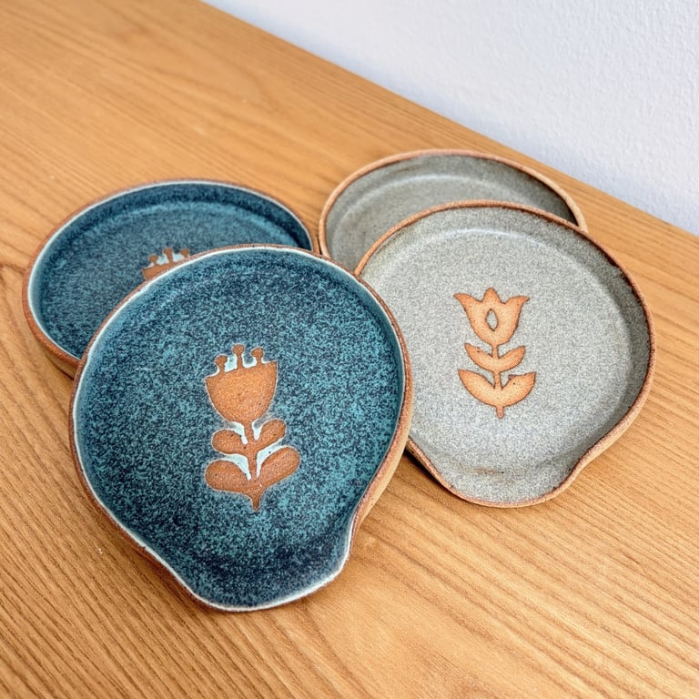 Fire små keramiske underkopper, to i blågrøn glasur og to i grå glasur, hver med et indgraveret blomsterdesign i midten, placeret på et lyst træbord.