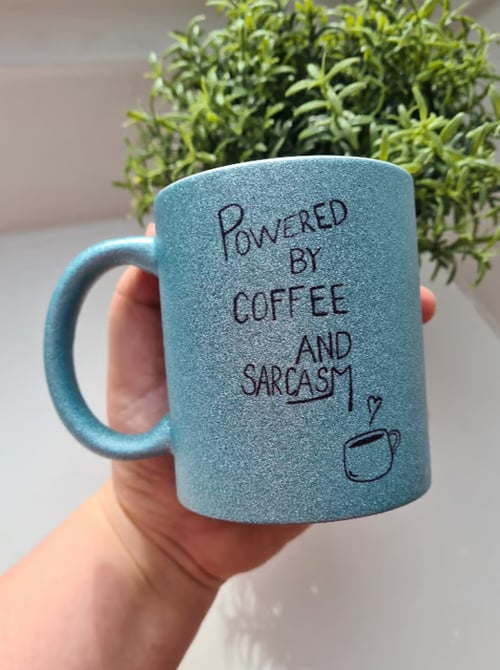 En hånd holder en blå glitrende krus med teksten "Powered by coffee and sarcasm" skrevet på. I baggrunden ses en grøn plante. (1)