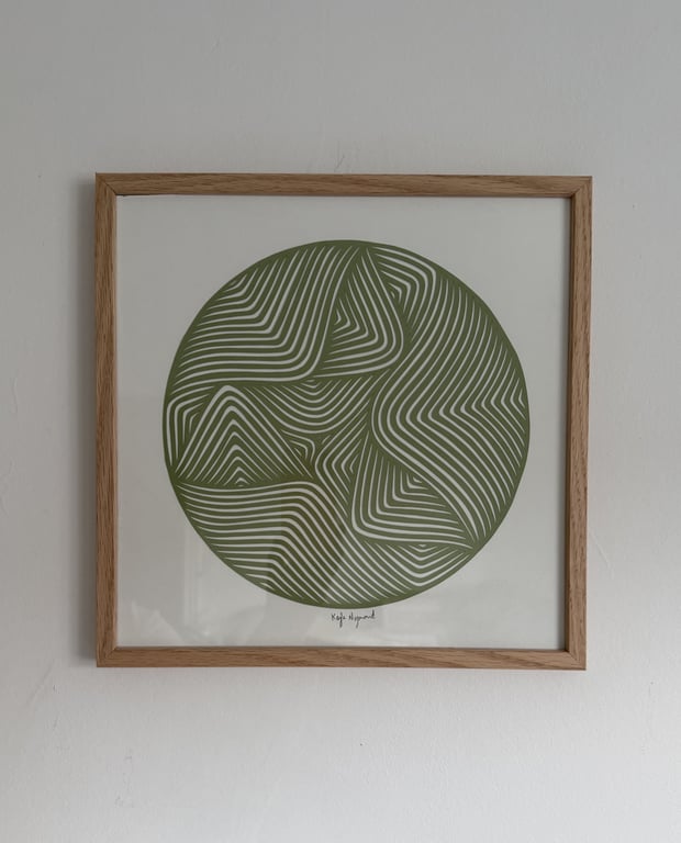 Abstrakt cirkelformet kunst med grønne og hvide bøjede linjer i et lyst træramme, hængende på en hvid væg.