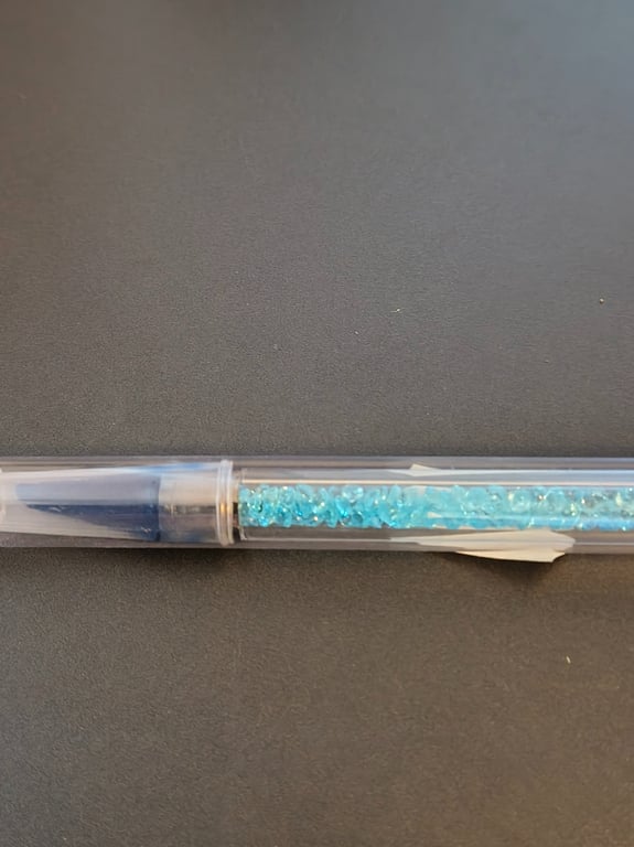 Kuglepen med gennemsigtig krop fyldt med små blå dekorative perler, placeret på en mørk overflade.