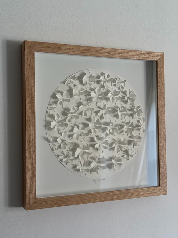Indrammet kunstværk med cirkel af hvide, tredimensionelle papirblomster på hvid baggrund og træramme.