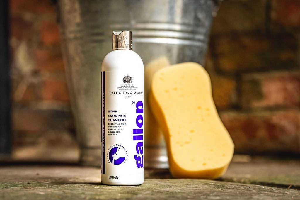 Flaske med Gallop pletfjerner shampoo og en gul svamp stående på en gulv med en metalspand og murstensvæg i baggrunden.