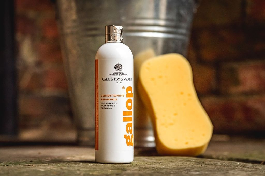 Flaske med Carr & Day & Martin Conditioning Shampoo Gallop ved siden af en gul svamp, med en metalspand i baggrunden.