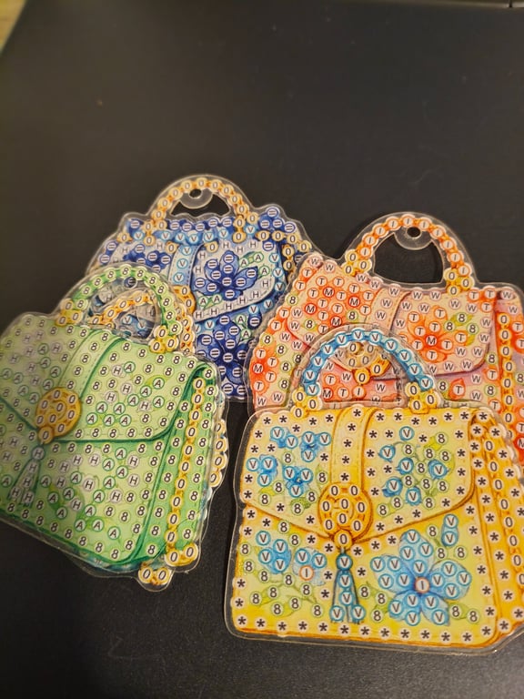 Fire farverige håndtasker lavet som broderi eller perleplade med bokstaver og symboler, placeret på en mørk overflade.