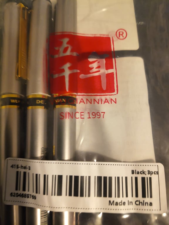 Billede af tre sorte penne i plastikemballage med mærkning "QIANNIAN SINCE 1997" og en stregkode med teksten "Made in China".