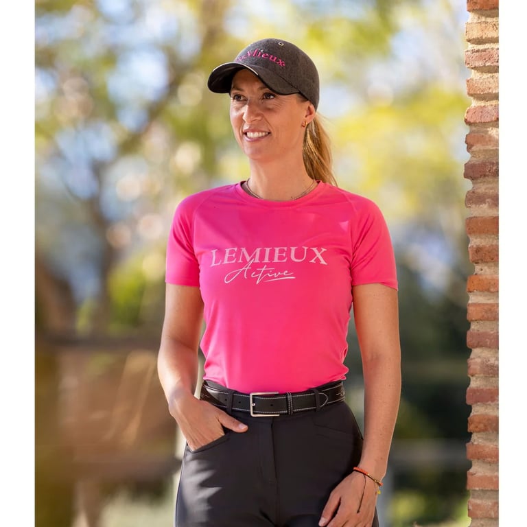 Kvinde iført pink Lemieux Active t-shirt og sort kasket, stående udendørs ved en mur med sløret naturbaggrund.