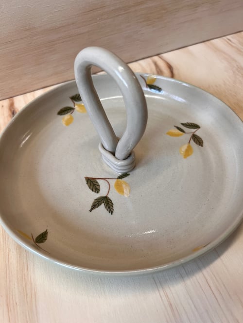 Keramisk fad med central hank og dekoration af små gule citroner med grønne blade på en beige baggrund. (1)
