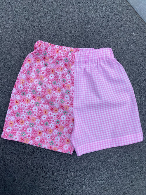 Lyserøde shorts med elastik i taljen, delt i to mønstre: blomsterprint på venstre side og lyserødt ginghamternet mønster på højre side. (1)