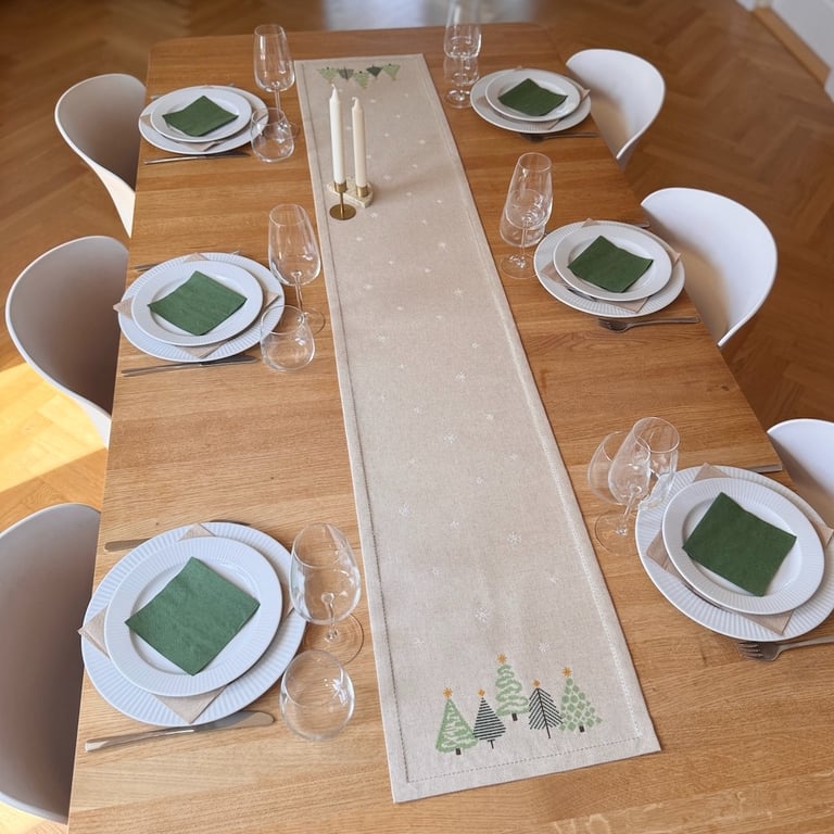 Spisebord med seks dækkede kuverter, hvide tallerkener, grønne servietter, vinglas og en beige bordløber med juletræsdekorationer og to hvide stearinlys i messingstager.