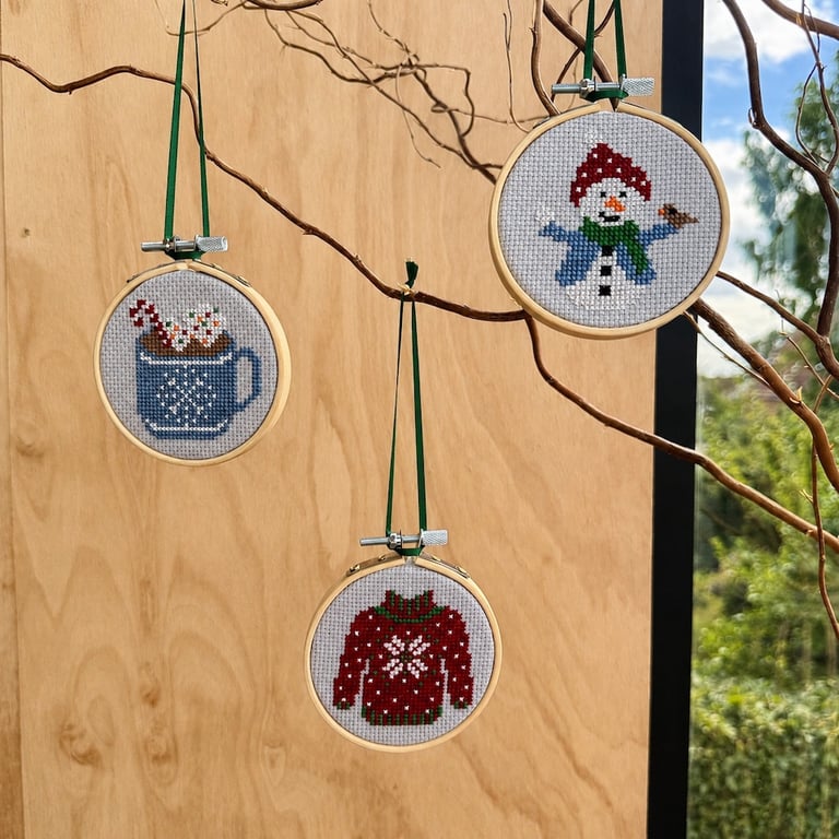 Broderede julemotiver med korssting i runde broderirammer hængende på grene: en kop med slikstokke, en snemand med rød hue og grøn tørklæde, samt en rød julesweater med hvidt mønster.