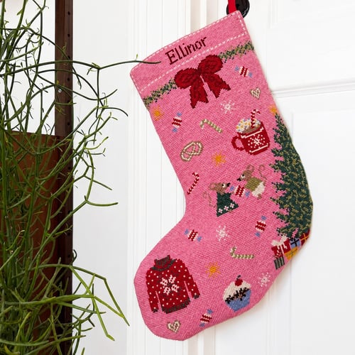 Julestrømpe i pink med broderede motiver som rød sløjfe, julesweater, mus, kaffekop, julekager og juletræ, hængende på en dør ved siden af en grøn plante. (1)