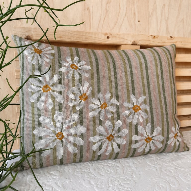 Rektangulær pude med stribede grønne og beige toner og hvide margueritblomster med gule centre, placeret på en lyse trækiste med grøn plante i venstre side.