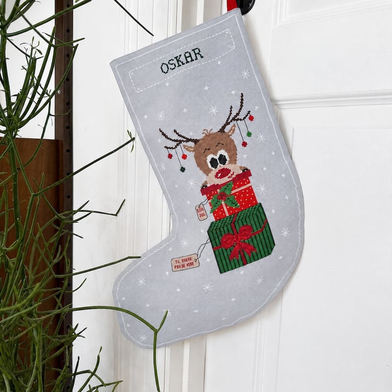 Julesok i gråt stof med broderet navn "OSKAR", der hænger på en hvid dør. Sokken er dekoreret med et broderet motiv af en rensdyrunge med julegaver og julekugler på gevir.