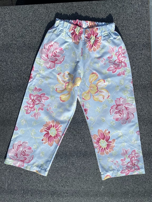 Blå bukser med elastik i taljen og blomsterprint i pink, gult og grønt, lagt fladt på en grå overflade.