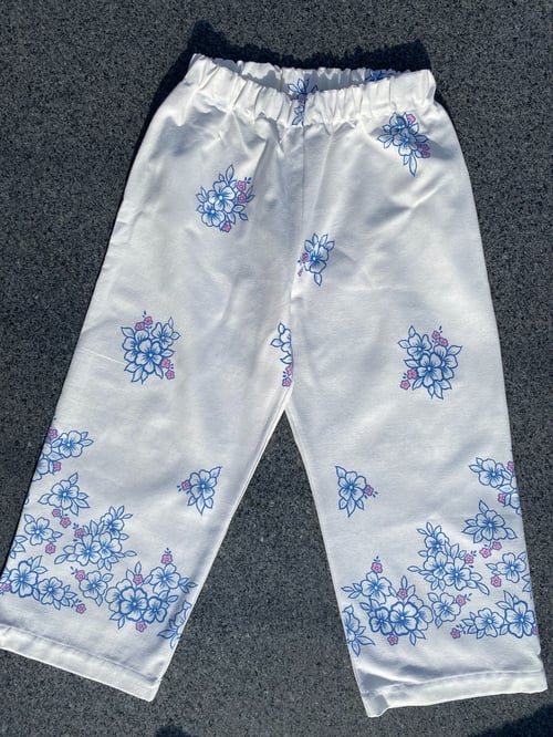 Billede af et par hvide bukser med elastisk talje og blå blomsterprint med små pink detaljer, lagt fladt på en mørk overflade. (1)