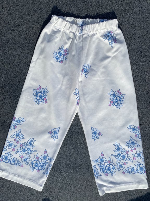 Billede af et par hvide bukser med elastisk talje og blå blomsterprint med små pink detaljer, lagt fladt på en mørk overflade.