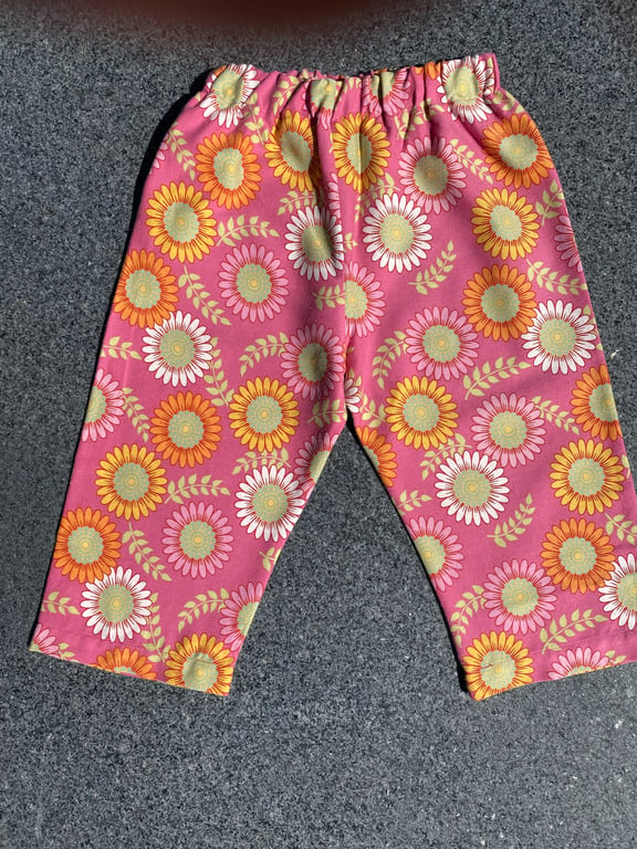 Bukser til børn med elastik i taljen, mønstret med store farverige blomster i orange, pink og hvid på pink baggrund, lagt fladt på mørkt underlag.
