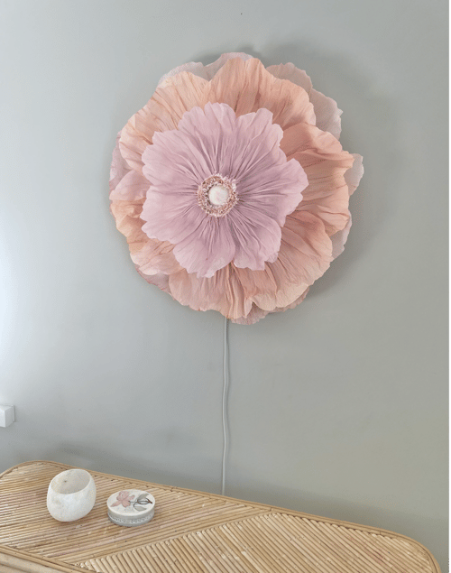 Stor lyserød og beige blomstervægdekoration monteret på grå væg over bambusbord med stearinlys og lille rundt låg. (1)