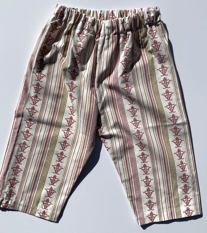 Et par løstsiddende bukser med elastisk talje, mønstret med lodrette striber og små røde symboler på beige bund.