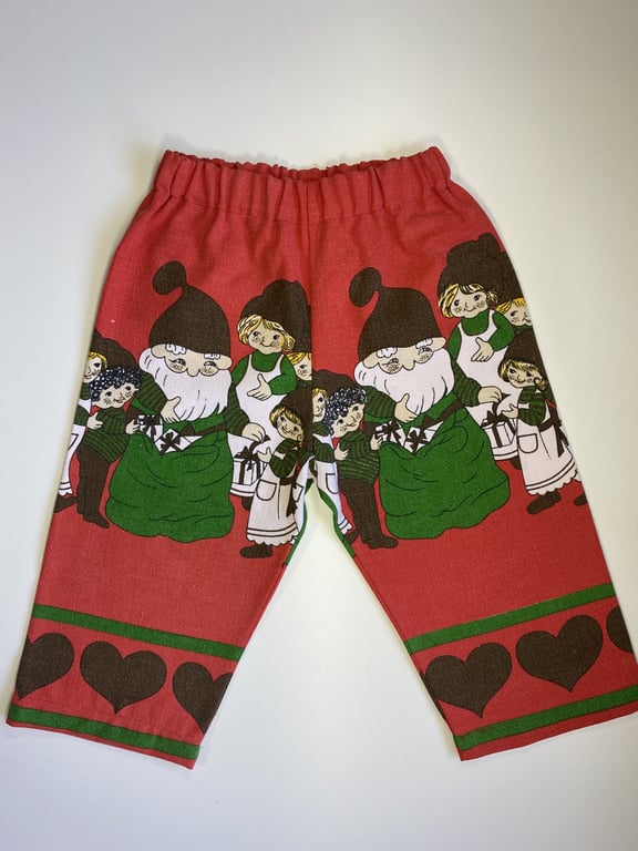 Bukser til børn med julemotiv, der viser nisser og piger i festlige farver rød, grøn og sort.