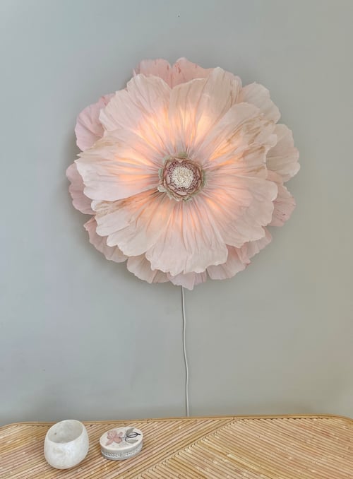 Væglys formet som en stor, lysende blomsterblomst i bløde lyserøde nuancer, monteret på en grå væg over et bord med to små dekorative genstande. (1)