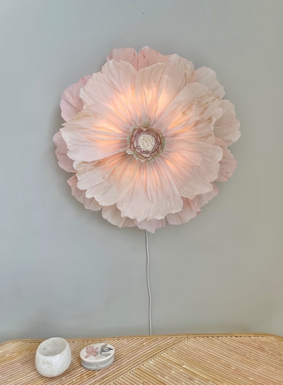Væglys formet som en stor, lysende blomsterblomst i bløde lyserøde nuancer, monteret på en grå væg over et bord med to små dekorative genstande.