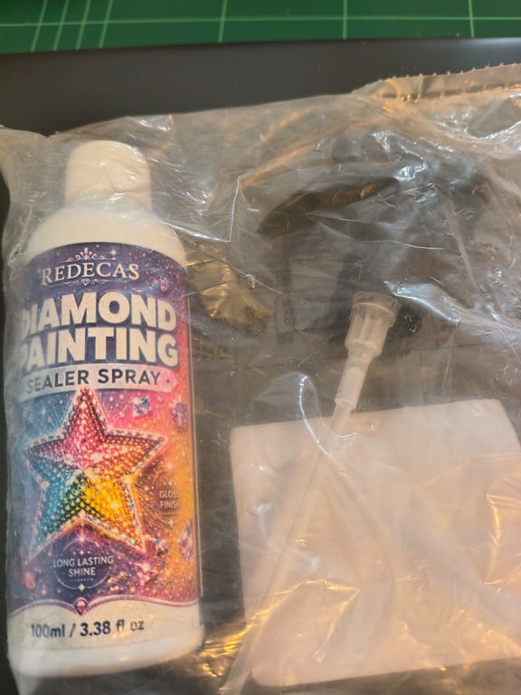 Flaske med Redecas Diamond Painting sealer spray og tilhørende sprøjte, begge pakket i en klar plastikpose.
