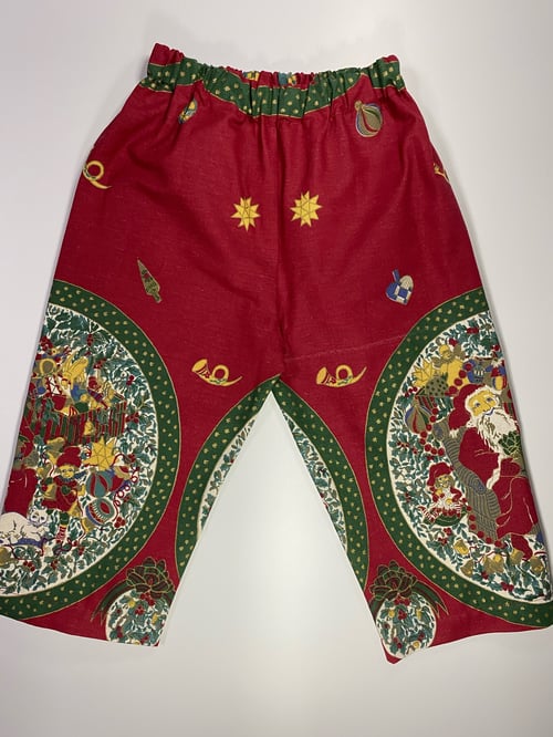 Bukser med juletema i rødt stof med grønne og gule motiver, herunder julemand, stjerner, kontrabas og julepynt. (1)