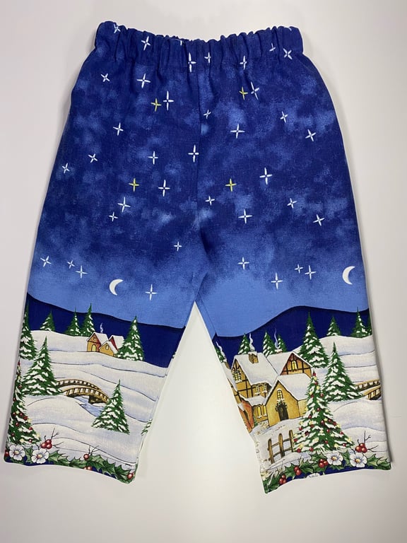 Børnebukser med julemotiv, der viser sneklædte landskaber, grantræer, små huse og en stjernehimmel med måner.