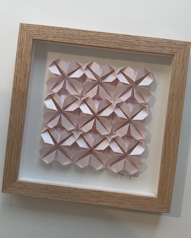 Indrammet kunstværk bestående af mønster af foldet, lyserødt papir arrangeret i et gentaget geometrisk design.