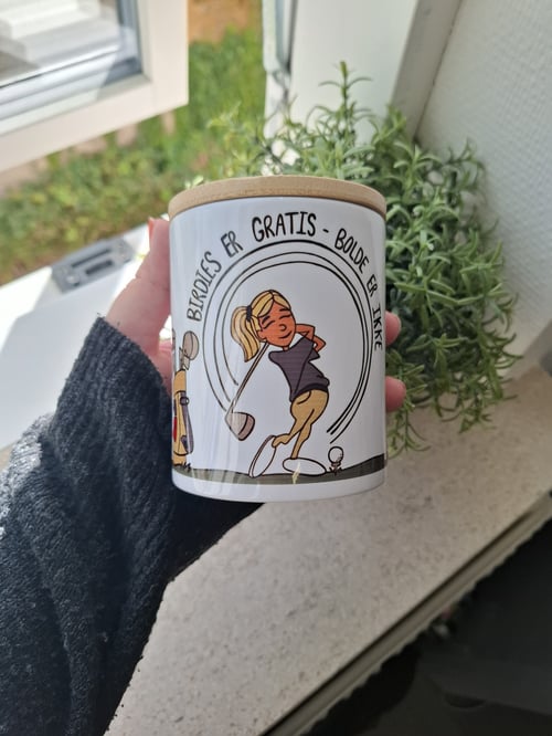 En hånd holder et krus med illustration af en kvinde, der spiller golf, og tekst på dansk, der siger: "Birdies er gratis - bolde er ikke". Baggrunden viser en vindueskarm med en grøn plante. (1)