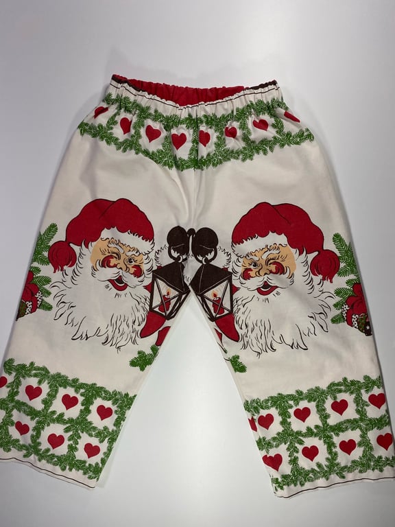 Bukser med julemotiv, herunder Santa Claus med rødt julehue, lanterner, grønne grangrene og røde hjerter på cremefarvet stof.