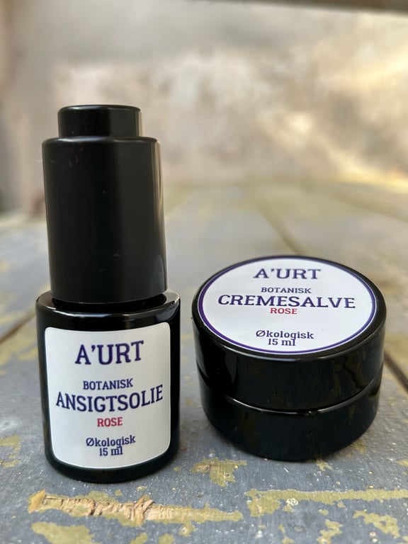 Sort flaske med botanisk ansigtsolie og sort krukke med botanisk cremesalve fra mærket A'URT, begge indeholdende 15 ml økologisk produkt med duft af rose, placeret på træbord.