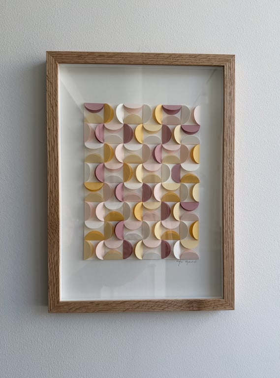 Indrammet moderne vægkunst bestående af tredimensionelle, buede geometriske former i pastelfarver beige, gul og rosa, arrangeret i et gentaget mønster mod hvid baggrund.