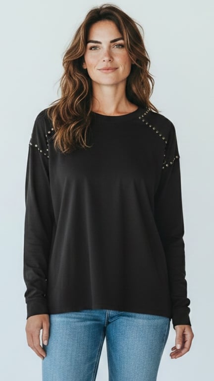 Kvinde iført sort langærmet bluse med metalpynt på skuldrene og lyseblå jeans, stående foran hvid baggrund.